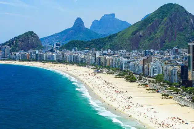 Orla de Copacabana lotada de pessoas aproveitando a praia, com o mar azul-turquesa e montanhas imponentes ao fundo.