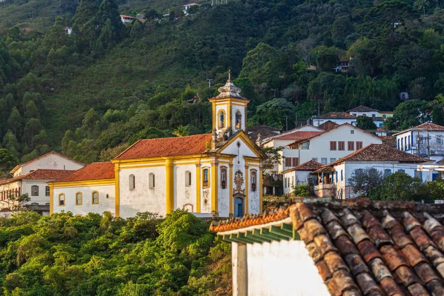 Igreja colonial branca e amarela de Nossa Senhora das Mercês e Misericórdia em Minas Gerais, com detalhes barrocos e paisagem natural ao redor.
