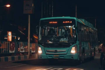 Um ônibus urbano de cor verde-água com as luzes acesas, parado em uma via noturna com sinalização de "STOP" visível no asfalto