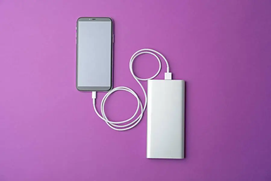 Celular conectado a uma bateria externa branca para recarga em superfície roxa.