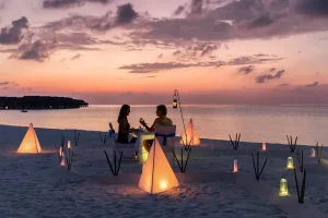 Casal jantando à luz de velas em praia paradisíaca ao entardecer.