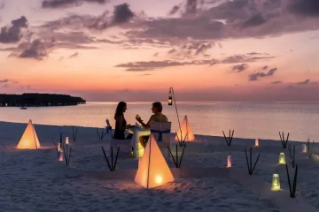 Casal jantando à luz de velas em praia paradisíaca ao entardecer.