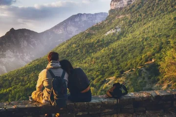 Casal viajante sentado em mureta de pedra observando paisagem montanhosa verdejante.