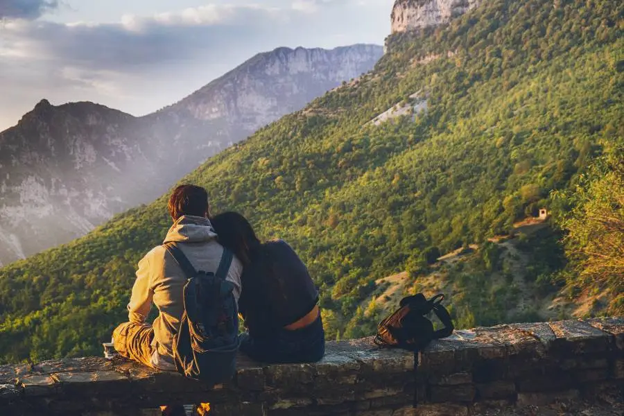 Casal viajante sentado em mureta de pedra observando paisagem montanhosa verdejante.